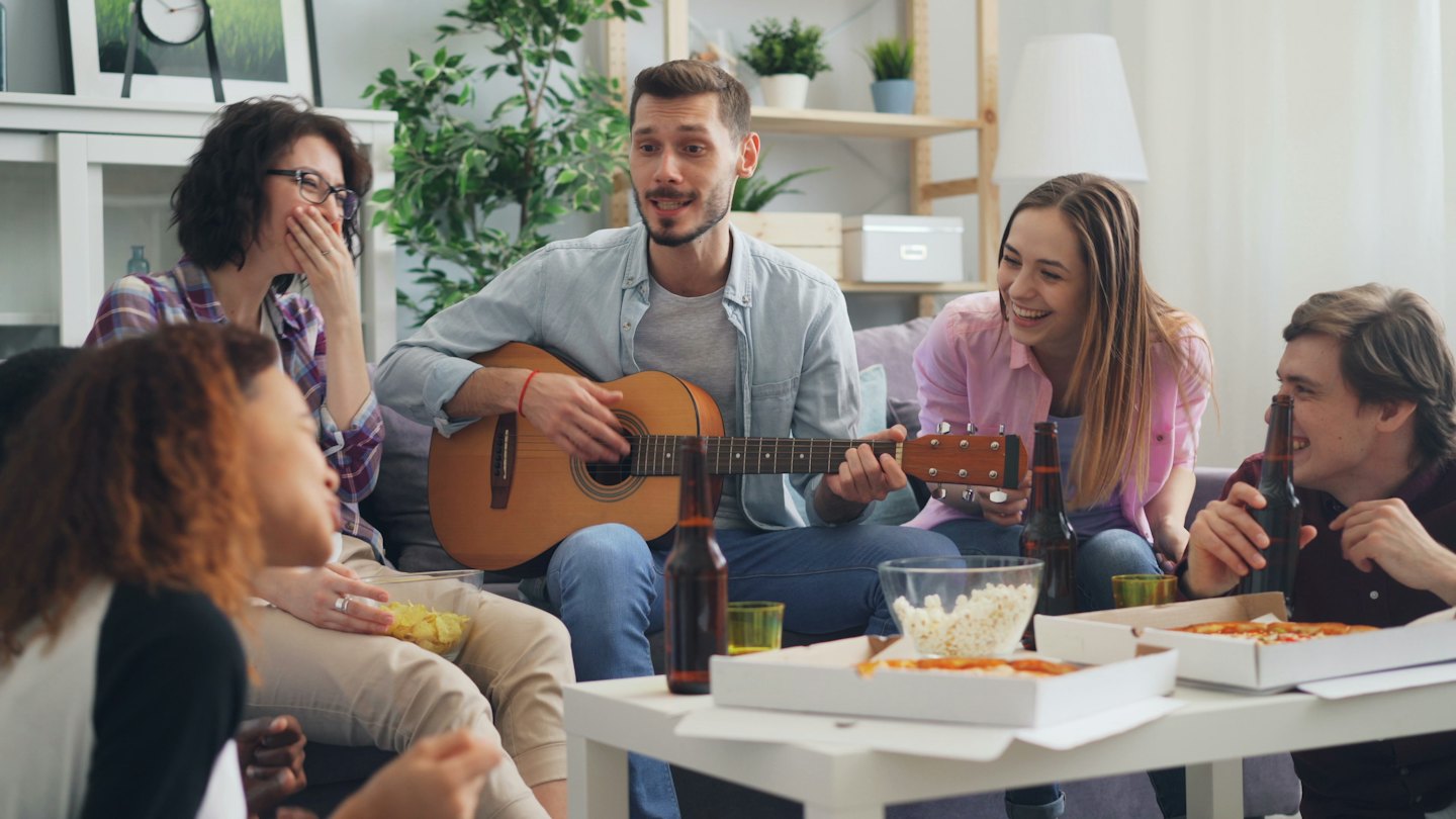 Five friends laugh while eating snacks and listening to guitar music.